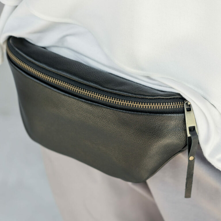 Close-up. A woman in a white blouse and gray pants wears a black leather banana bag
